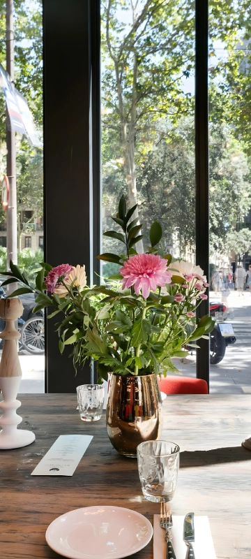 Arreglo de flores rosadas en florero dorado junto a una mesa, cerca de una ventana