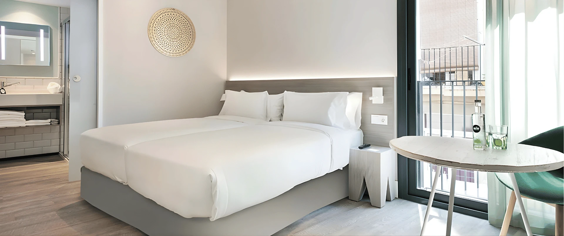 Habitaci&oacute;n de hotel moderna con cama blanca, mesa y luz natural por ventana