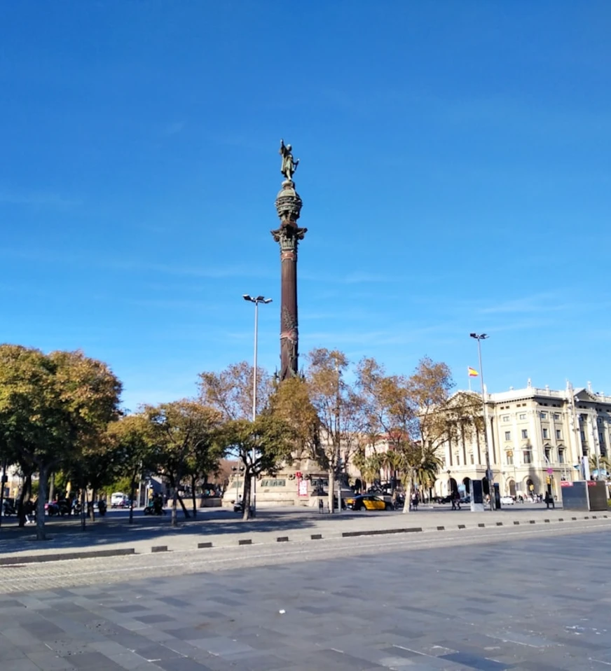 Columna con estatua y palmeras frente a edificios en una plaza, cielo azul