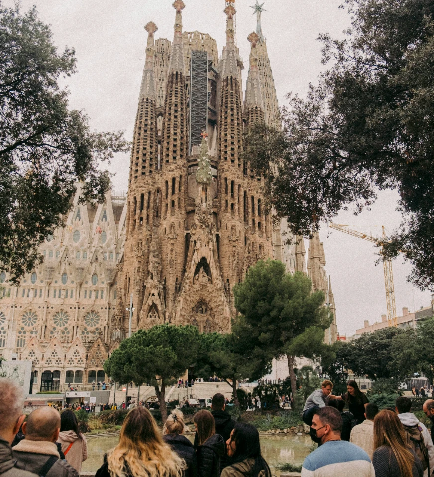 Sagrada Fam&iacute;lia en Barcelona, vista desde el jard&iacute;n con gente y &aacute;rboles alrededor