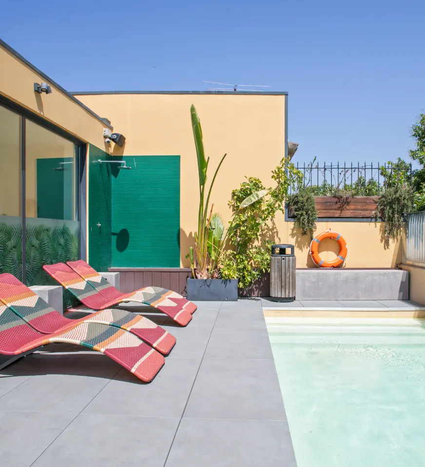 Piscina con tumbonas rojas y verdes, plantas y un edificio al fondo
