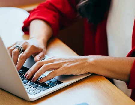 Persona con chaqueta roja escribiendo en una laptop sobre un escritorio.