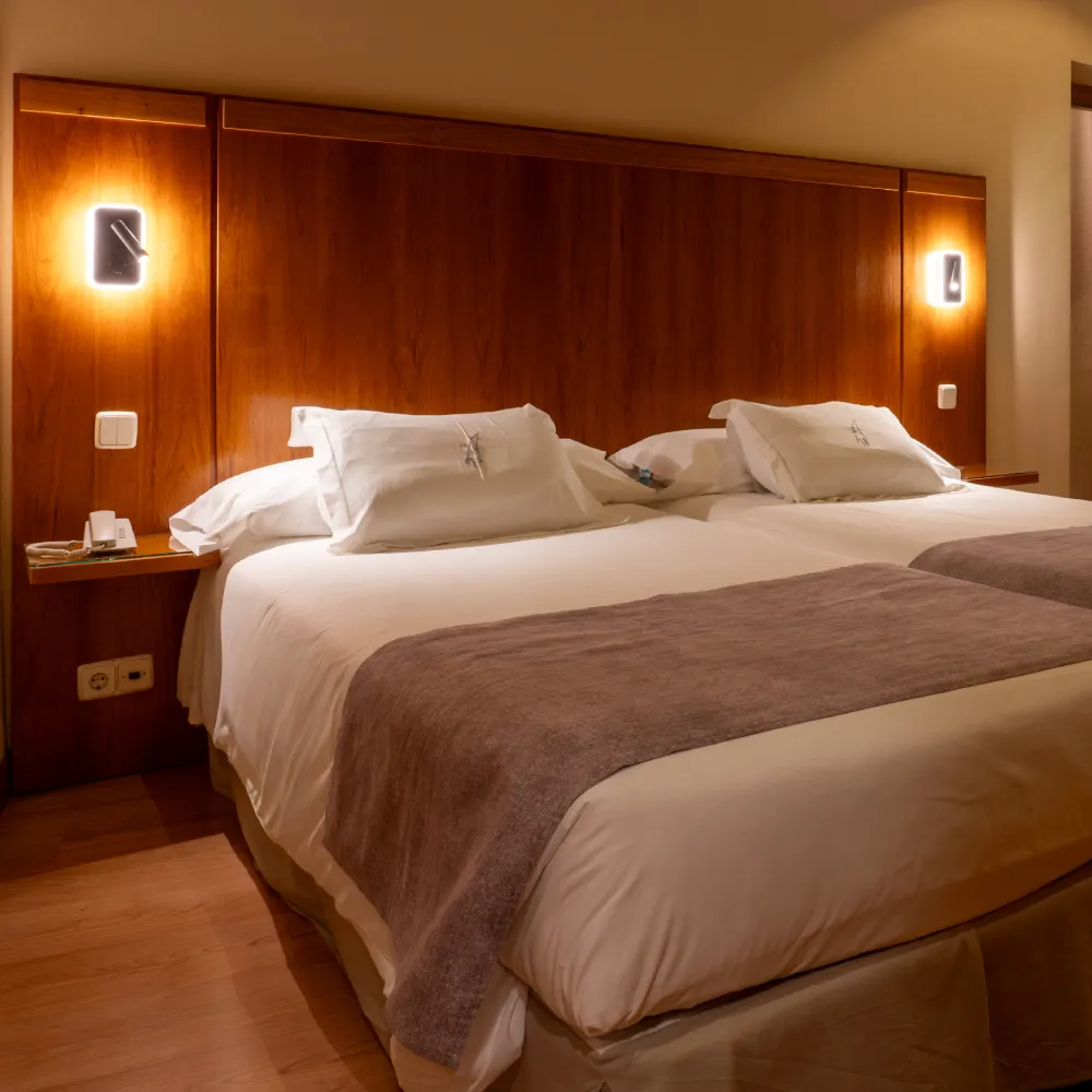 Cama doble en habitaci&oacute;n de hotel con cabecero de madera y l&aacute;mparas encendidas.