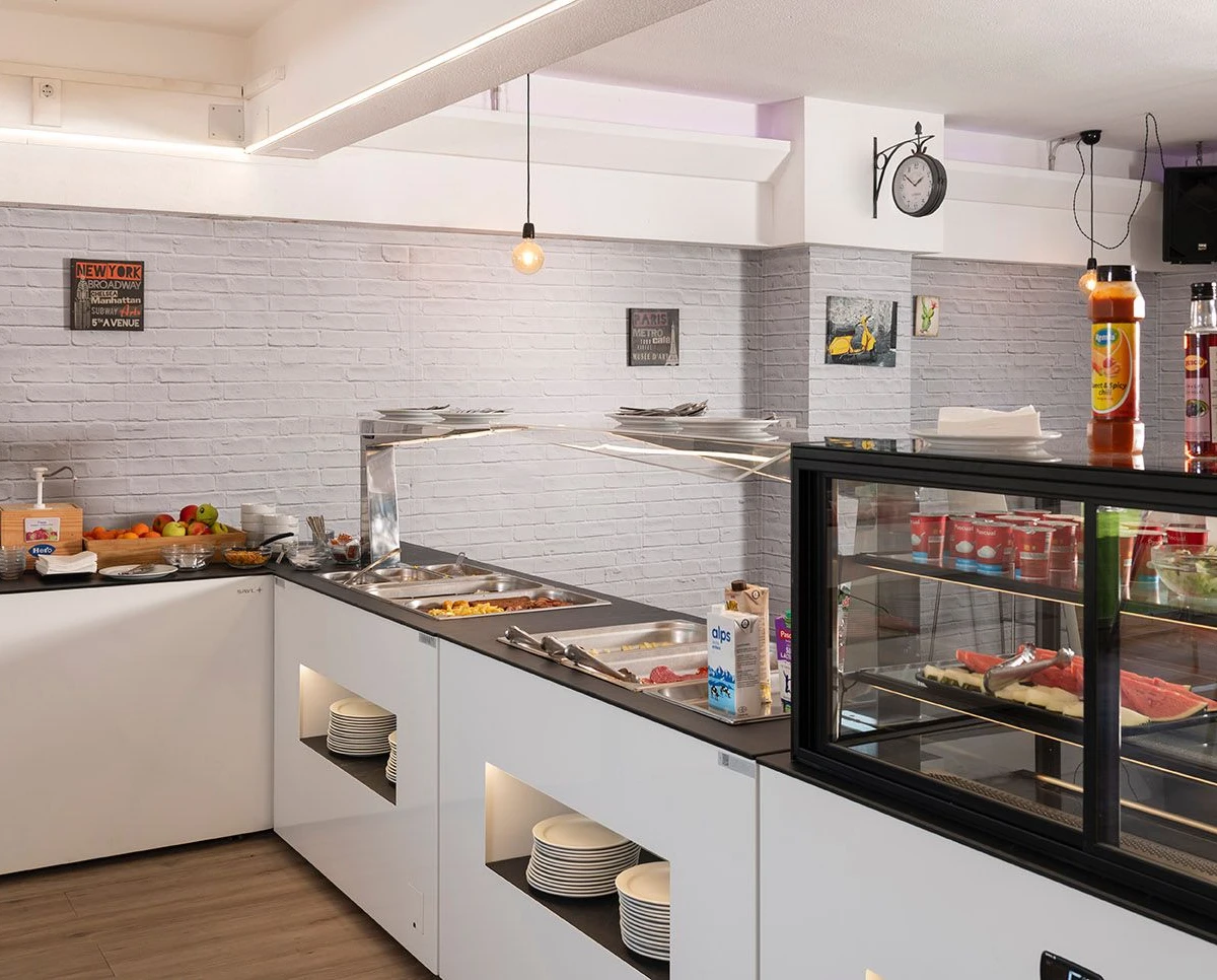 Cafetería con barra blanca, vitrinas y buffet con comida.