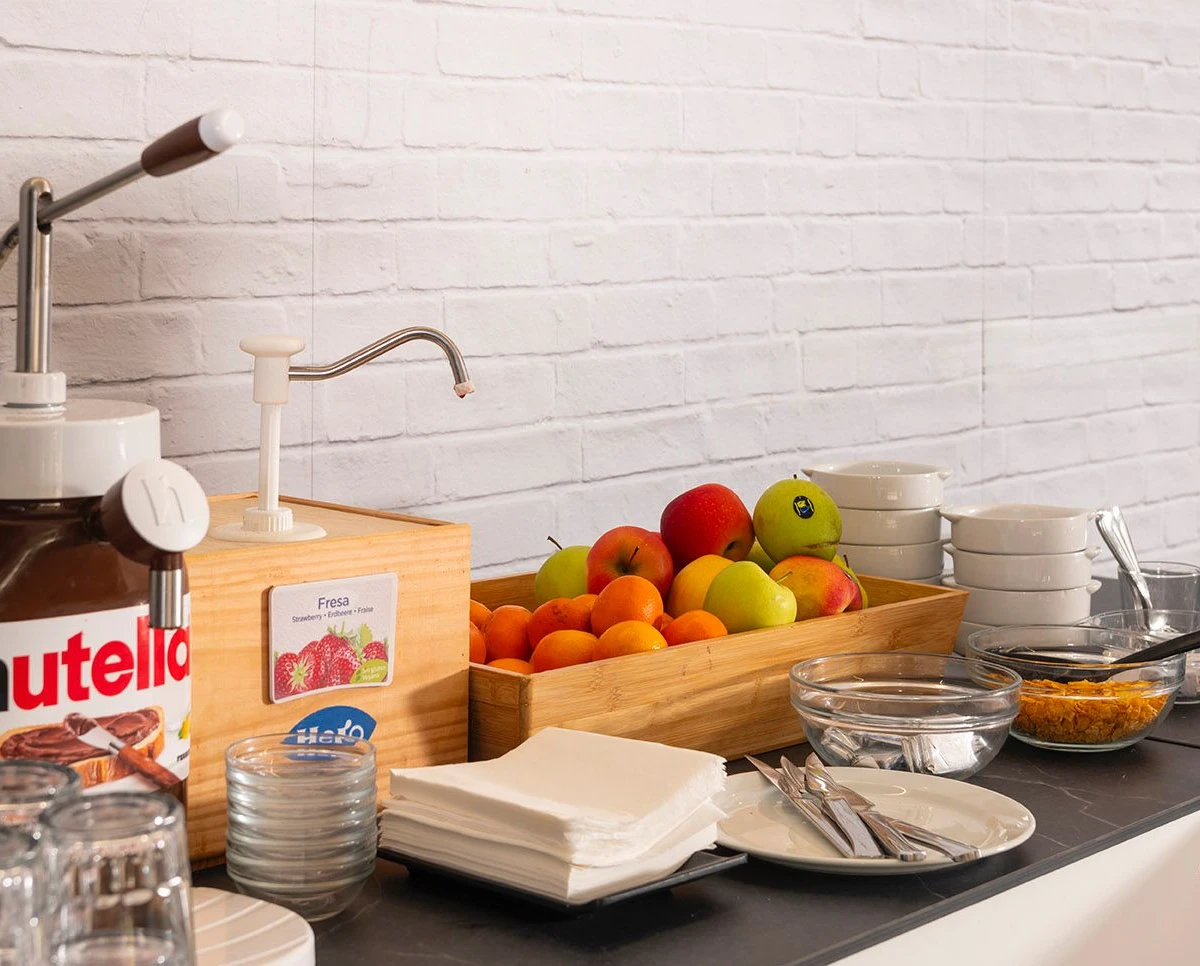 Mesa de desayuno con frutas, dispensador de chocolate y recipientes de comida