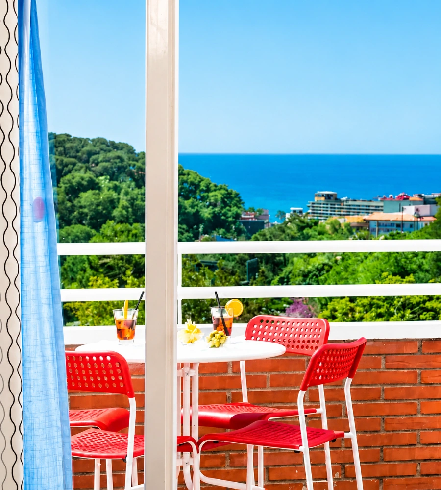 Mesas y sillas rojas en terraza con vista al mar y vegetaci&oacute;n, con cortina azul