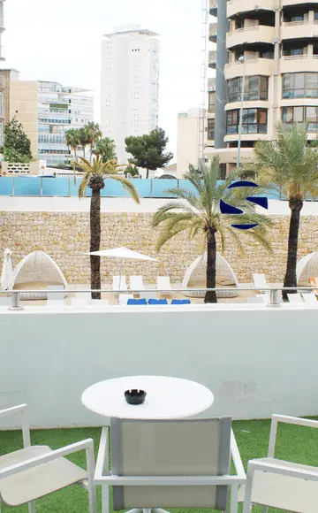 Terraza de hotel con vista a piscina y palmeras, silla y mesa blanca.