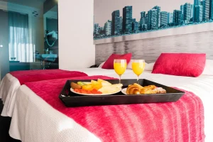 Habitación de hotel con cama roja, bandeja con desayuno y dos copas amarillas