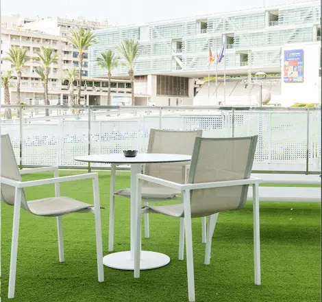 Terraza con mesas y sillas sobre c&eacute;sped, palmeras y edificio moderno al fondo