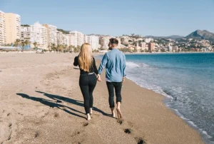 Pareja caminando de la mano por la playa con edificios y monta&ntilde;as al fondo.