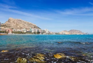 Vista costera con mar azul y edificios frente a montañas y rocas.