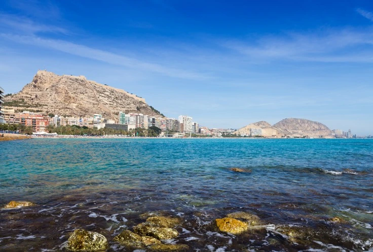 Vista costera con mar azul y edificios frente a monta&ntilde;as y rocas.