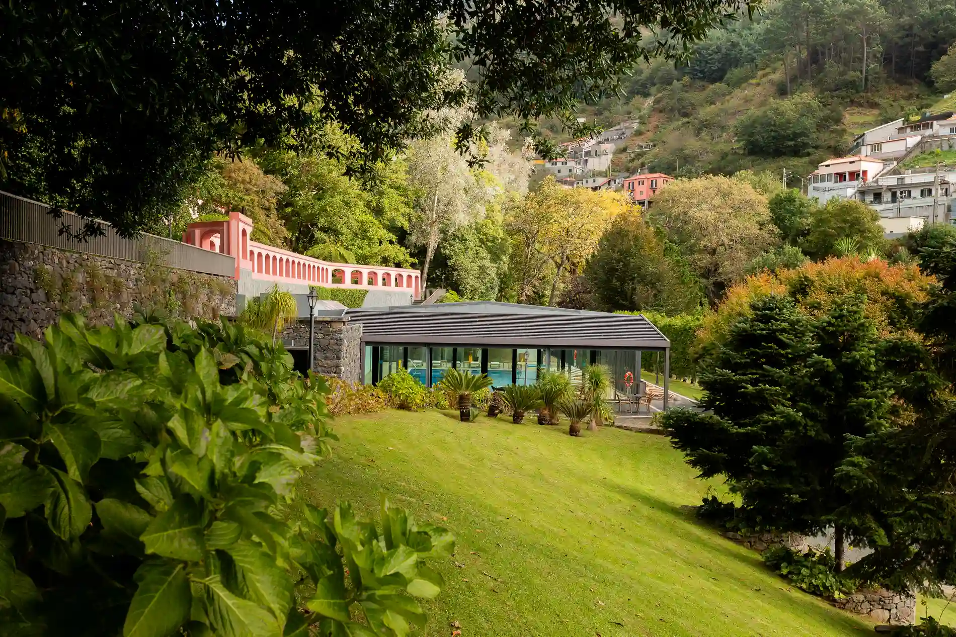 Jardim com edif&iacute;cio moderno rodeado de &aacute;rvores e vegeta&ccedil;&atilde;o densa.