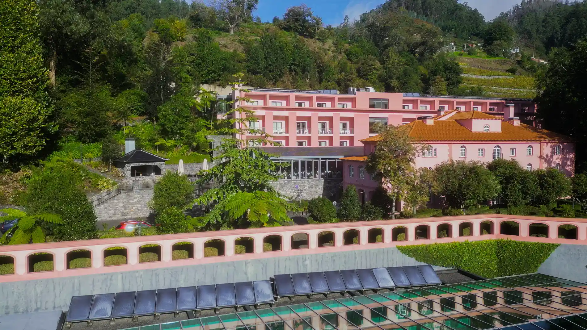 Pr&eacute;dio rosa cercado por vegeta&ccedil;&atilde;o e telhados alaranjados ao fundo.