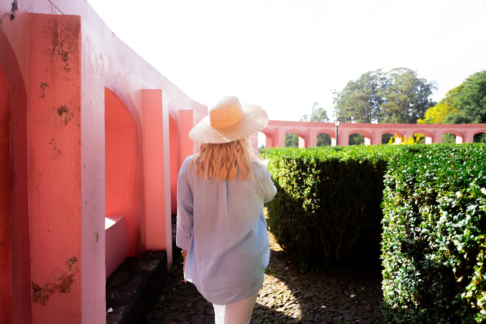 Mulher de chap&eacute;u andando em um jardim com paredes rosas.