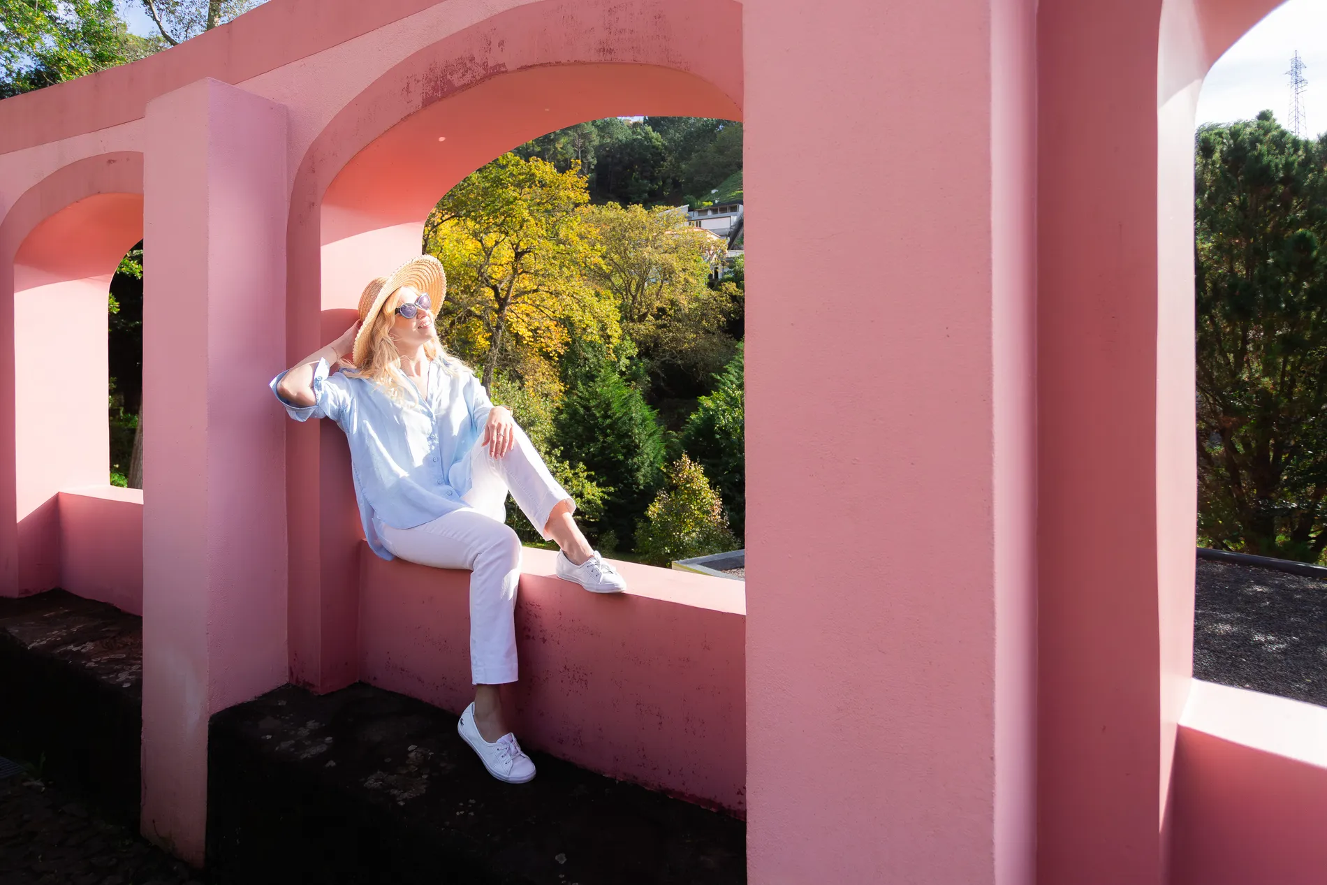 Mulher em roupa clara e chap&eacute;u, sentada em muralha rosa sob o sol.