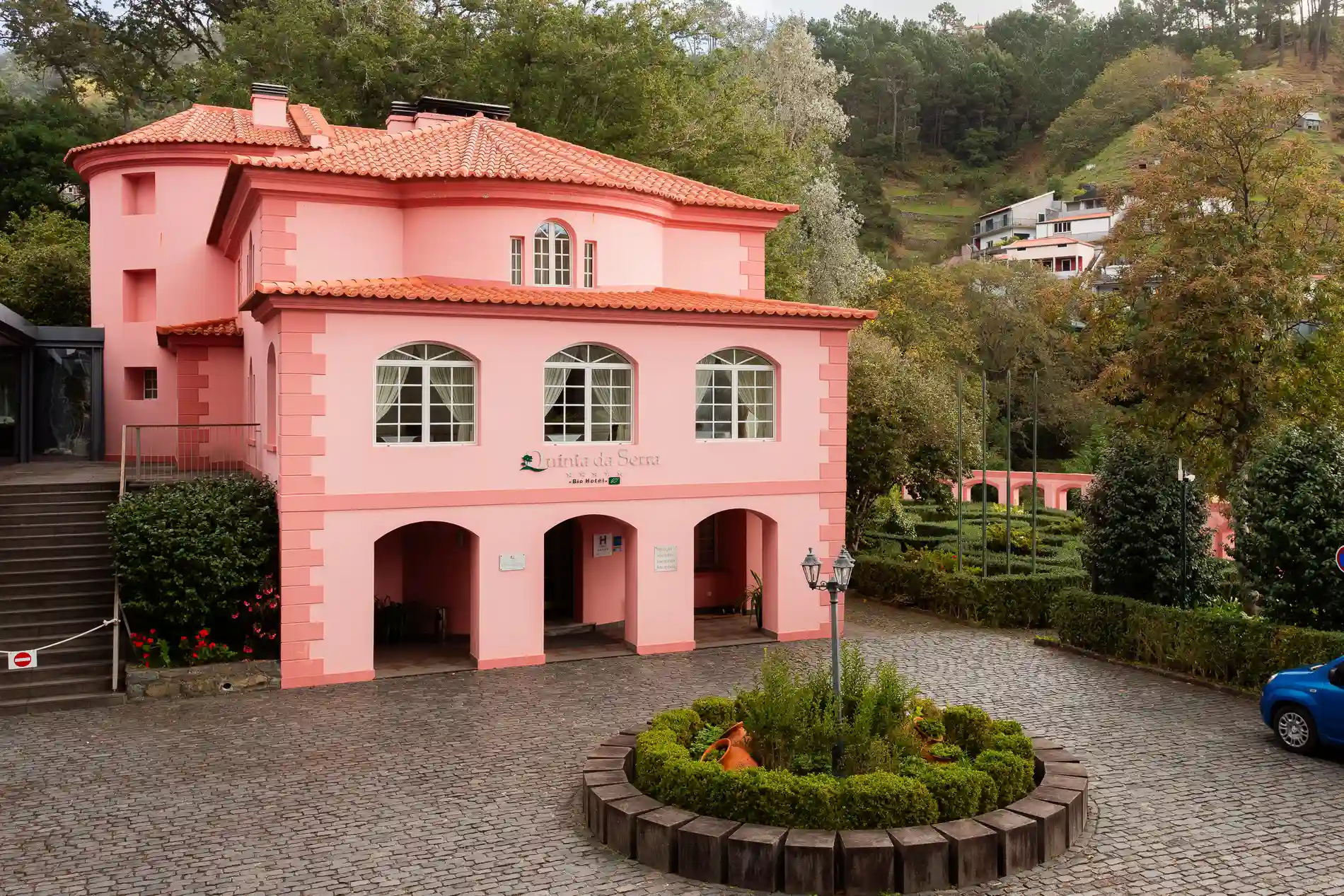 Edif&iacute;cio rosa em &aacute;rea arborizada com jardim e carro azul estacionado.