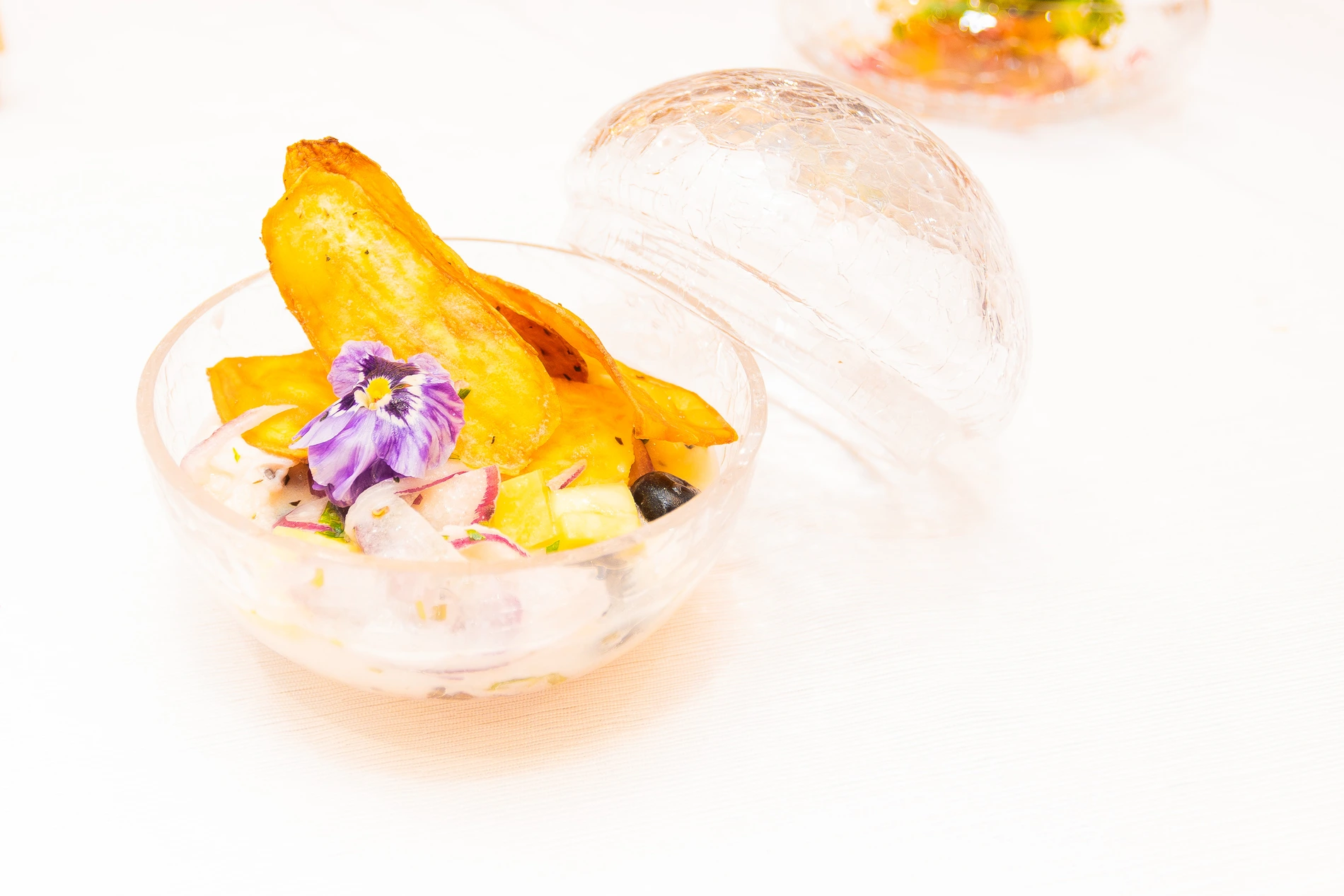 Tigela de vidro com ceviche decorado com flor roxa e batata-doce crocante.