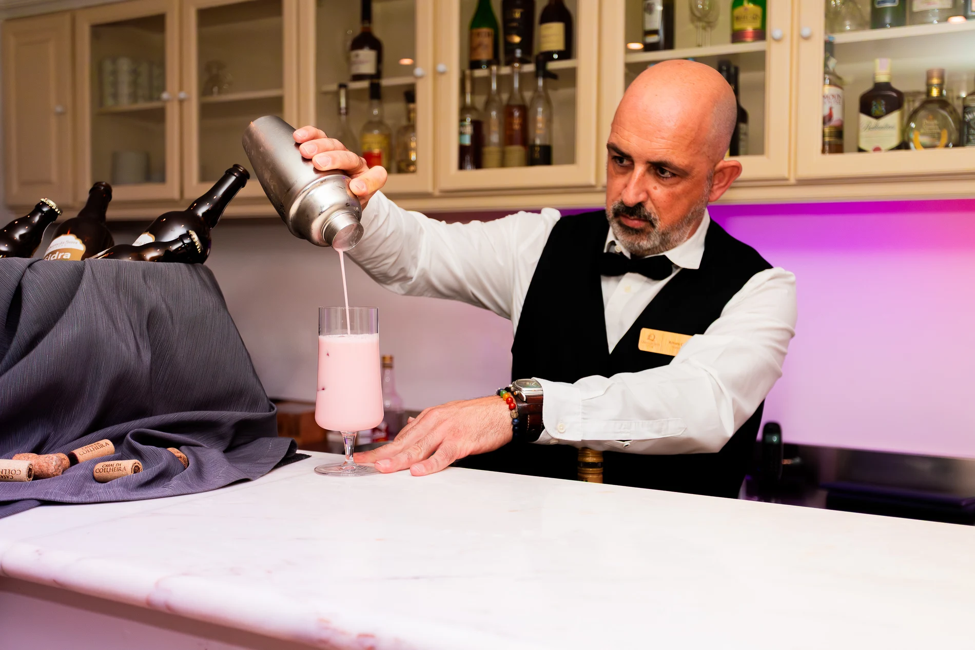 Bartender preparando drink rosa no balc&atilde;o de bar com garrafas ao fundo.