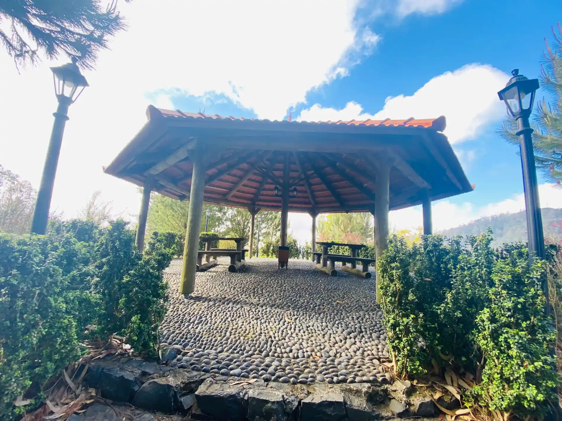 Quiosque de madeira sob c&eacute;u azul com vegeta&ccedil;&atilde;o ao redor.