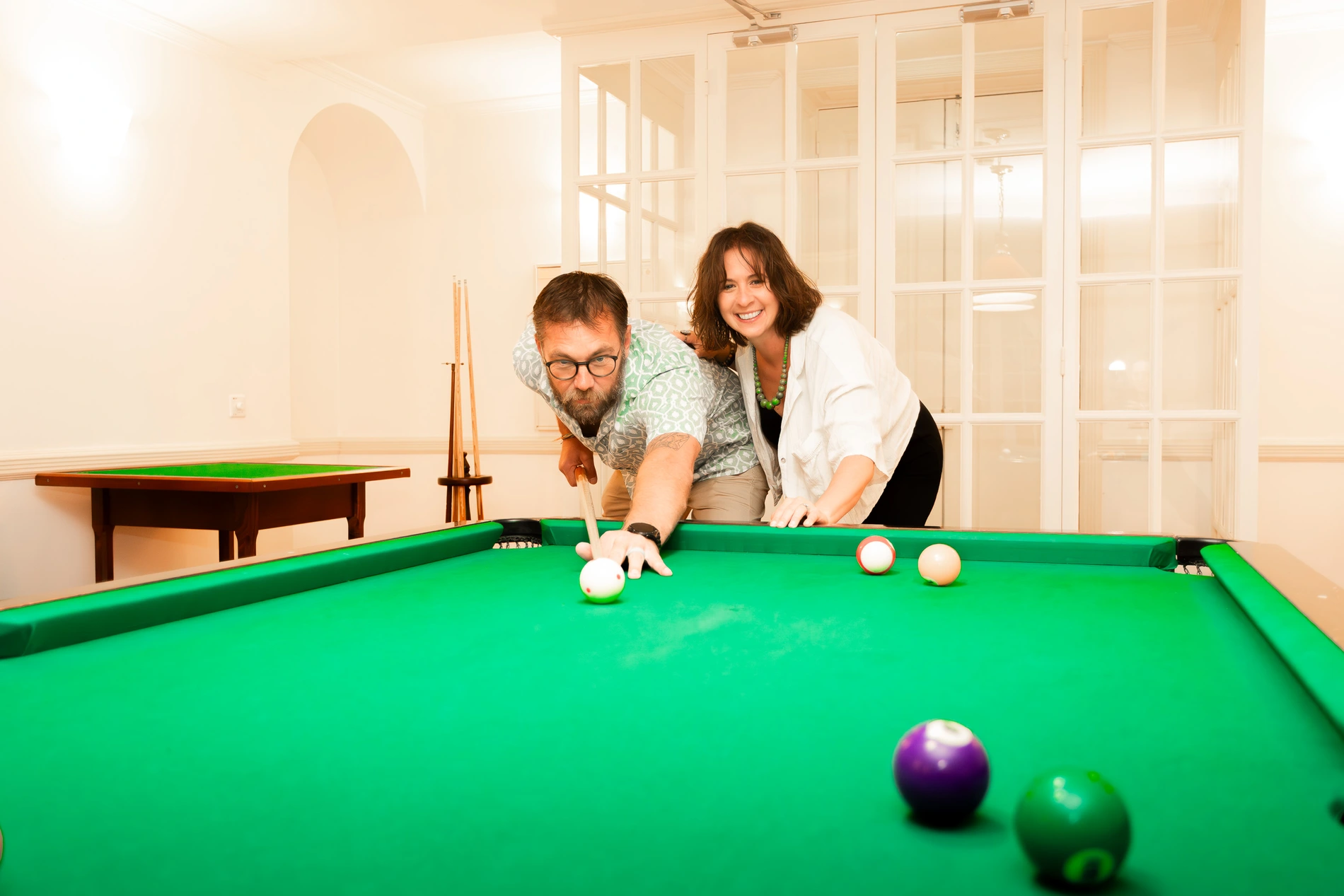 Casal jogando sinuca com bolas coloridas em uma sala bem iluminada.