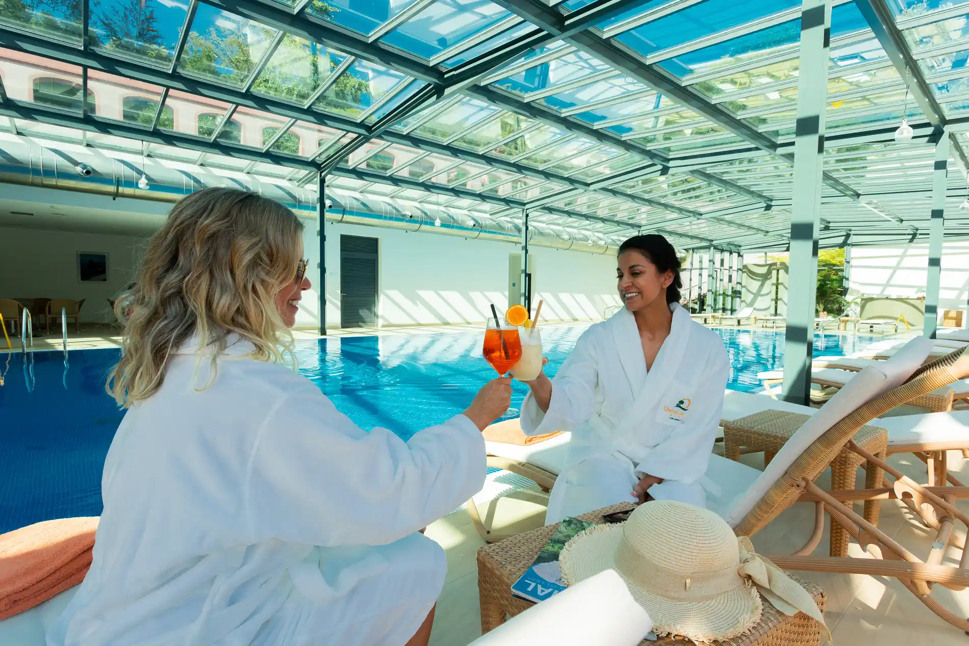 Duas pessoas brindam com drinques ao lado de uma piscina coberta.