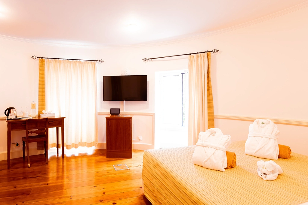 Quarto de hotel aconchegante com mesa, TV e roup&otilde;es na cama.