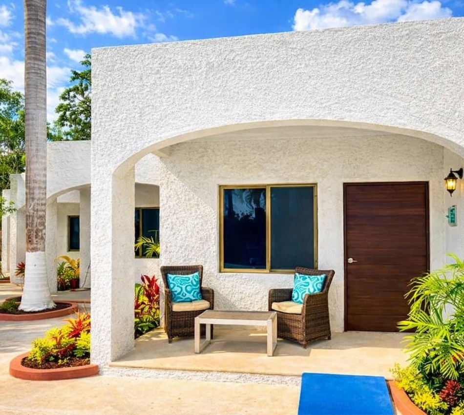 Casa blanca con sillas en el porche rodeada de plantas y cielo azul.