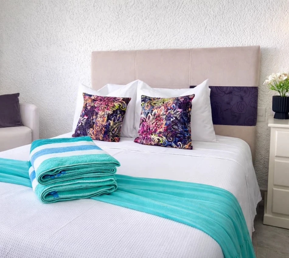 Cama de hotel con cojines coloridos y toallas dobladas azul turquesa.
