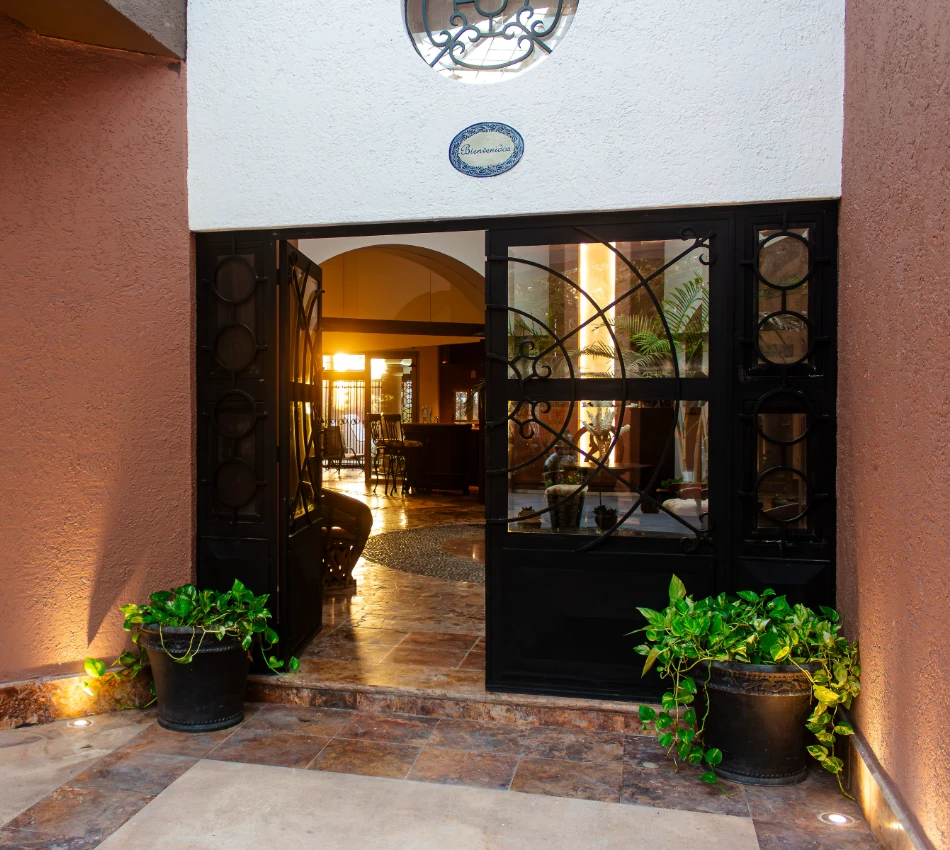 Entrada de casa con puerta abierta, plantas y luces c&aacute;lidas en el interior.