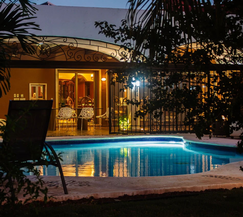 Piscina iluminada frente a una casa con terraza al atardecer.