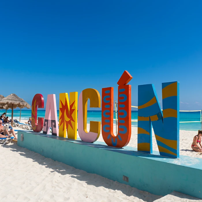 Letrero colorido de "CANC&Uacute;N" en la playa con cielo azul y sombrillas.