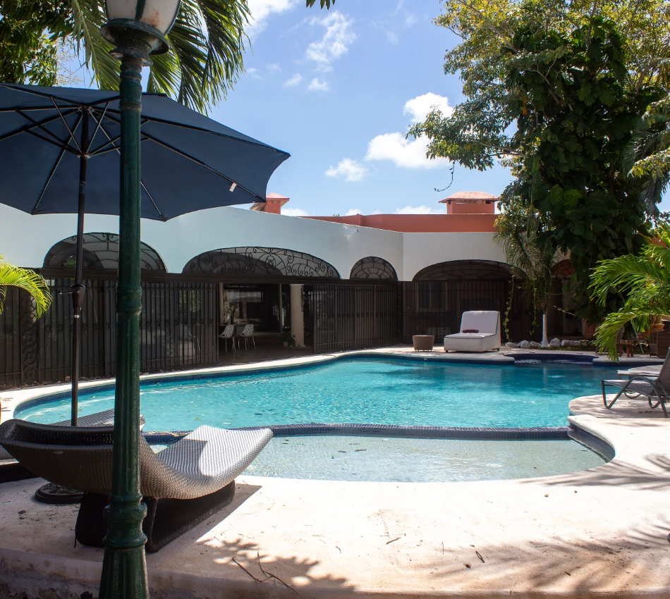 Piscina con sombrilla y muebles rodeada de vegetaci&oacute;n y casa de fondo.