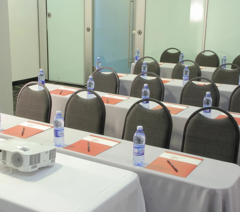 Sala de reuniones con sillas, mesas, agua, cuadernos y proyector.