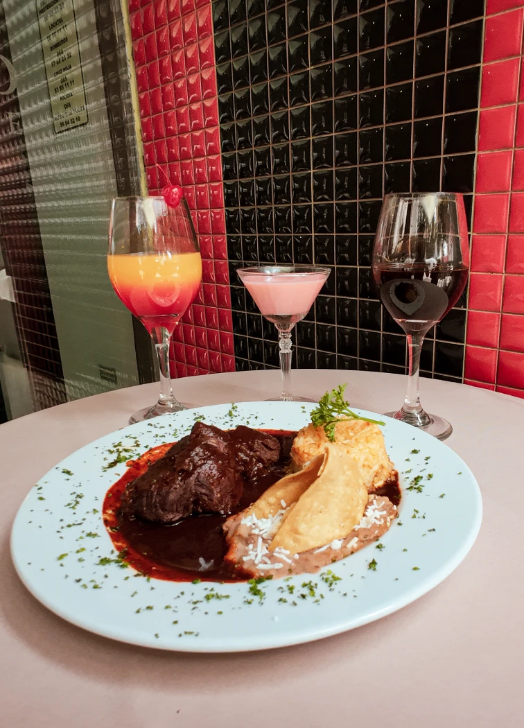Plato de mole con bebidas variadas en una mesa, fondo de mosaicos rojos y negros.