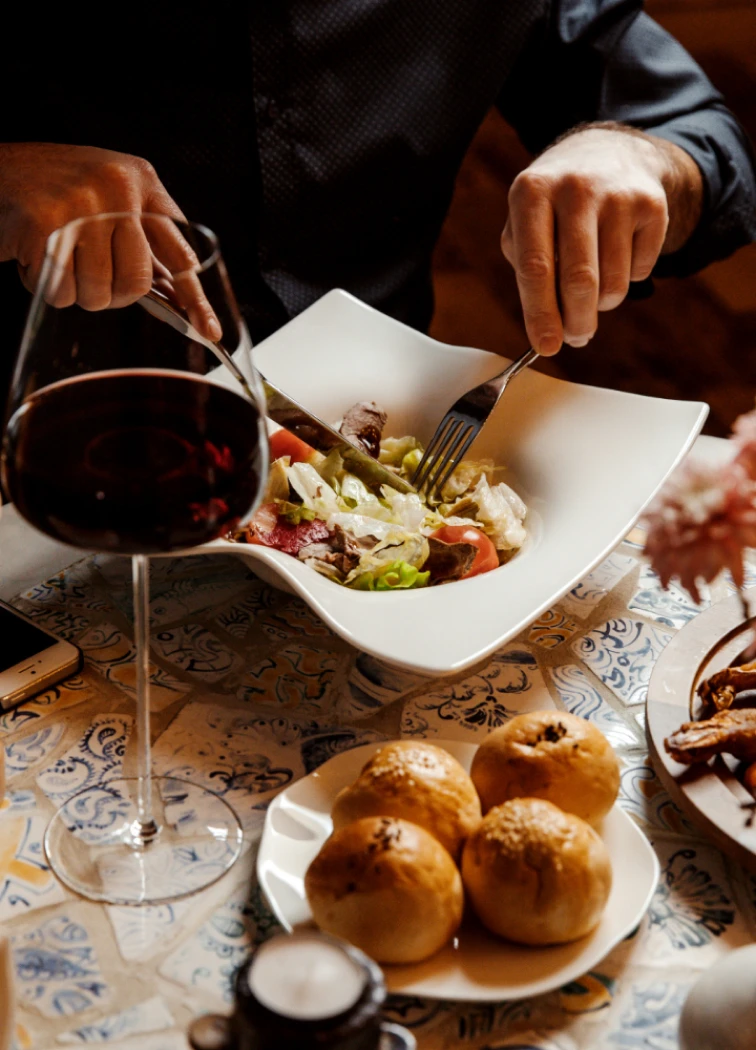 Persona comiendo ensalada con vino tinto y bollos sobre la mesa.