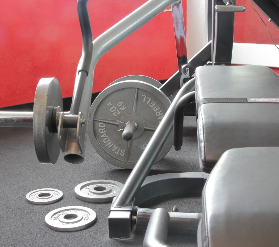 Pesas y banca de gimnasio sobre una alfombra negra, fondo rojo.