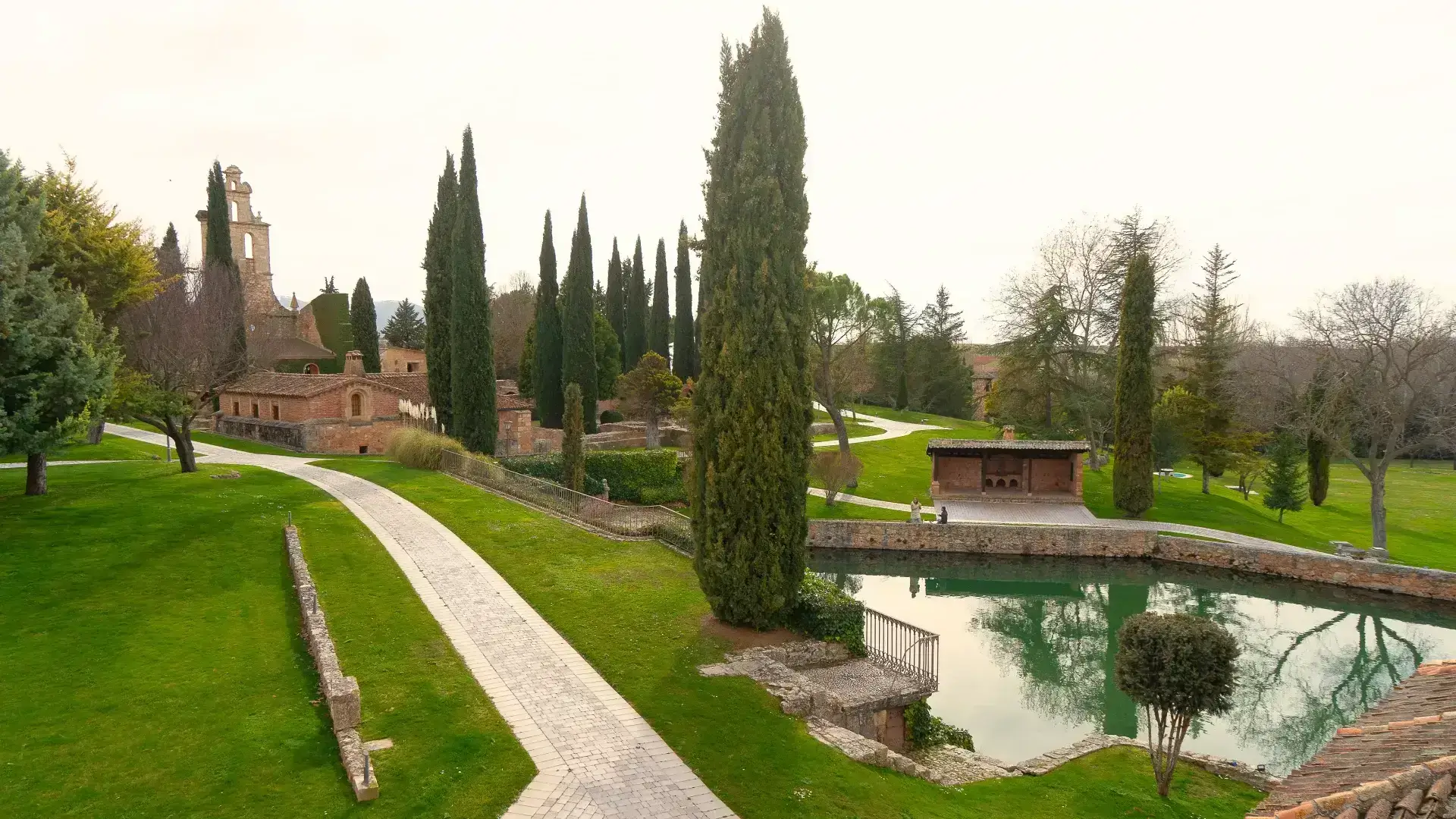 Jard&iacute;n con sendero, cipreses y estanque reflejando &aacute;rboles.