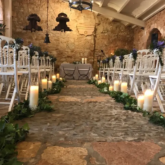 Pasillo de boda r&uacute;stico con velas y flores en un sal&oacute;n de piedra.