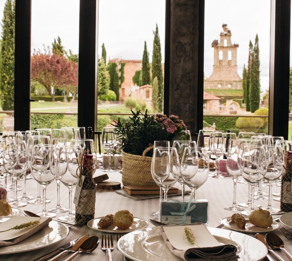 Mesa elegante con copas listas, vista exterior de jard&iacute;n y edificio antiguo.