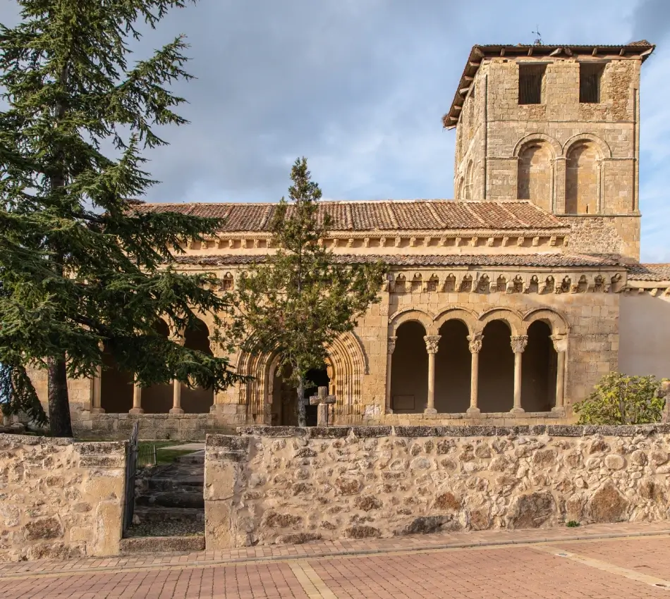 Iglesia rom&aacute;nica con arcos y campanario de piedra, rodeada de &aacute;rboles.
