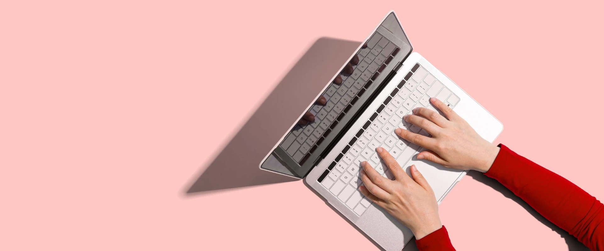Manos escribiendo en una laptop sobre un fondo rosa.