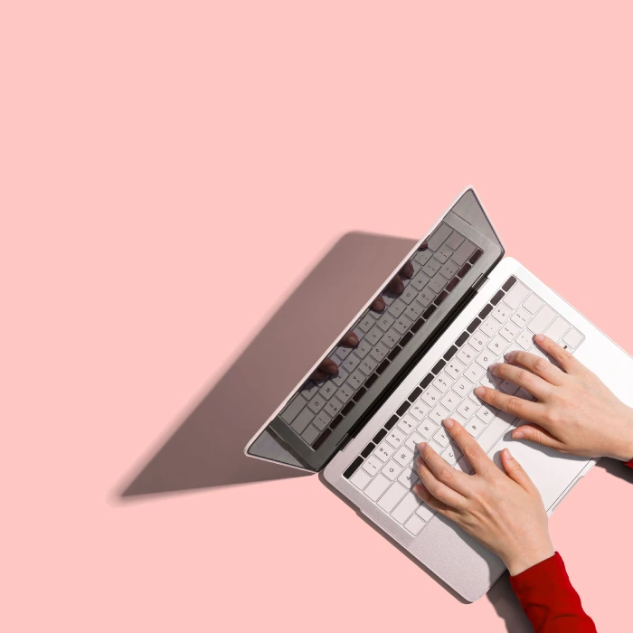 Manos sobre un teclado de laptop plateada sobre fondo rosa.