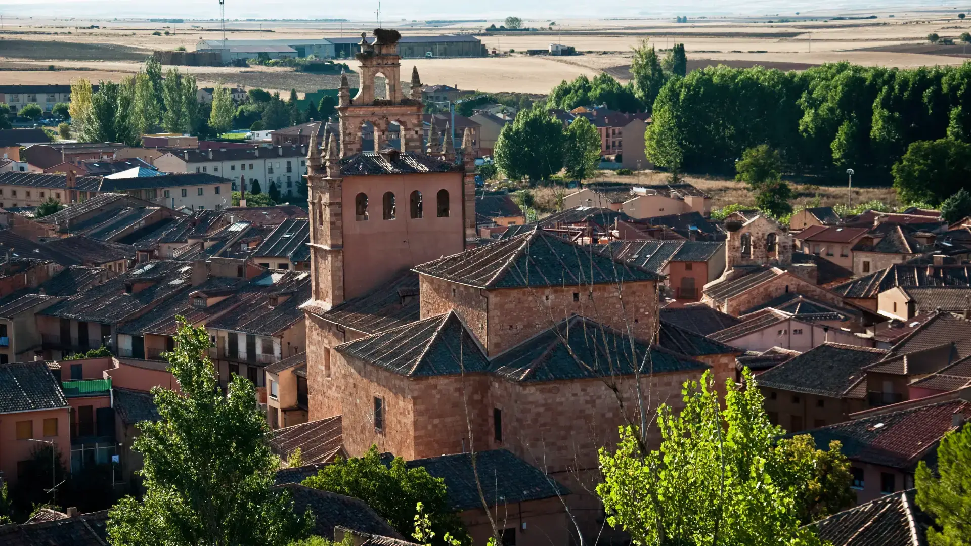 Pueblo con iglesia de piedra y techos de teja rodeado de &aacute;rboles y campos.