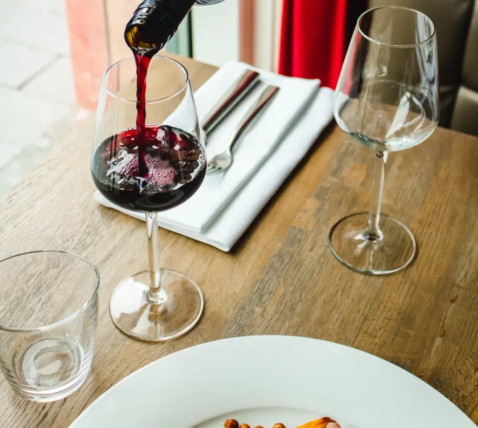 Vino tinto sirvi&eacute;ndose en copa junto a plato y cubiertos sobre mesa de madera.