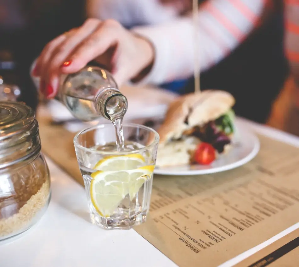 Mano sirviendo agua en vaso con lim&oacute;n junto a un plato con pan y ensalada.