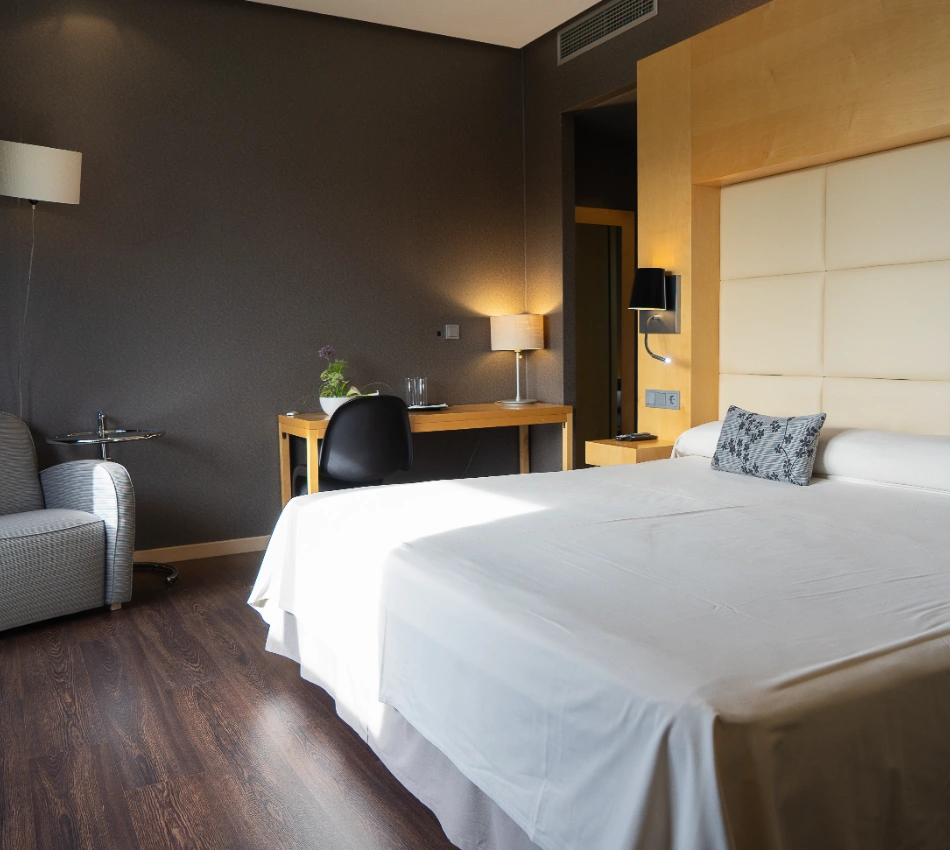 Habitaci&oacute;n de hotel moderna con cama, escritorio y sill&oacute;n gris.