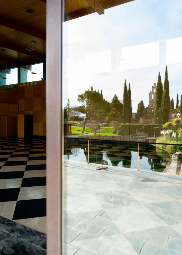Reflejo de un jard&iacute;n y edificios en un vidrio junto a suelo ajedrezado interior.