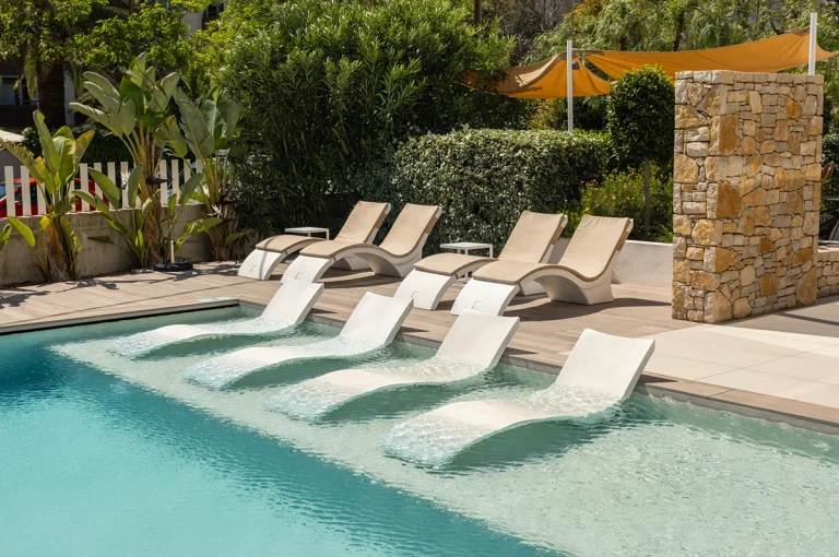 Piscina moderna con tumbonas y vegetación de fondo en un día soleado.