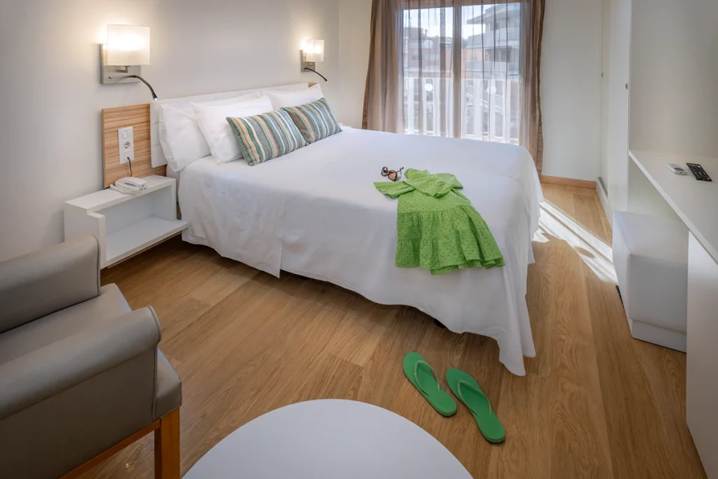 Cama matriominio con cojines en habitaci&oacute;n iluminada, mesas y sillas.